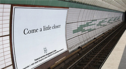 Large white poster that reads Come a little closer is affixed to the wall of an underground train station, opposite the platform and across the tracks.